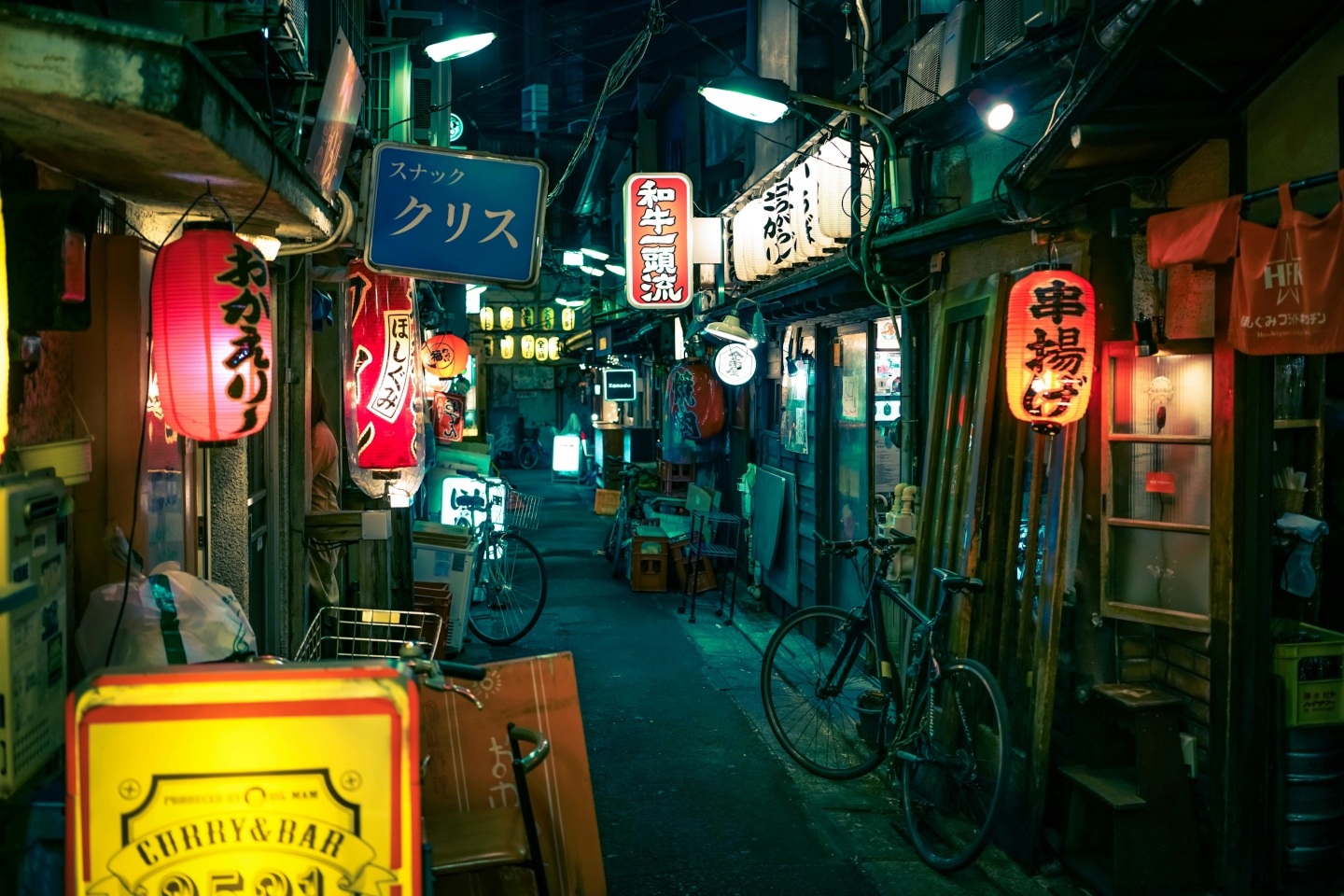 A narrow alleyway of shops and restaurants in Japan with some signs, lights, and a couple bicycles in frame, at night