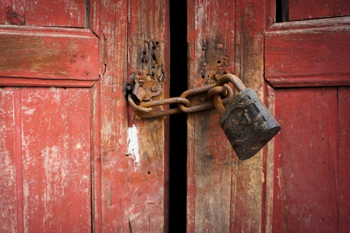 Two wooden doors with red paint are slightly ajar, opening inward, but held together with a rusty chain and padlock.